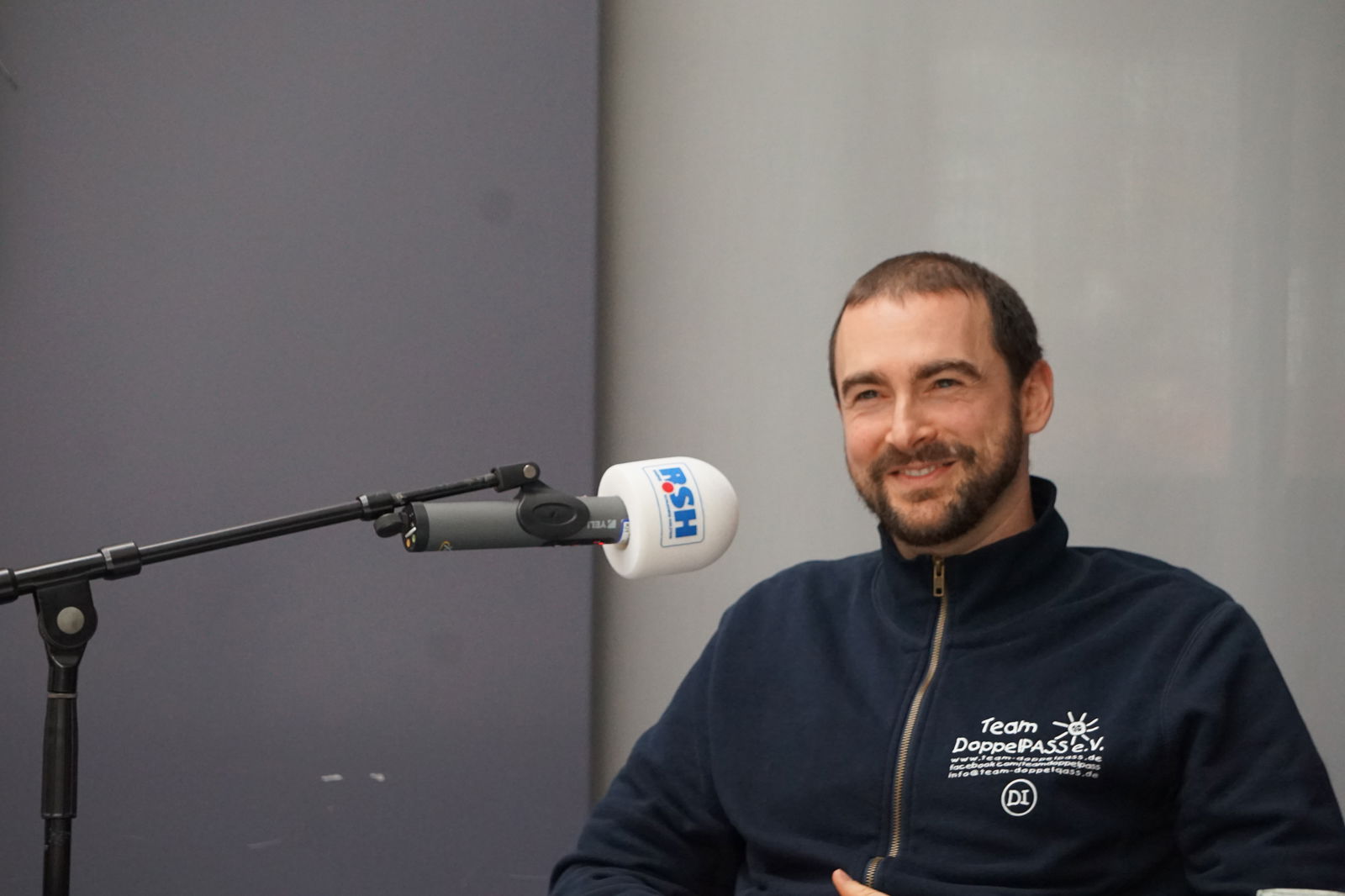 Ein Mann mit kurzen Haaren sitzt auf einem Stuhl und spricht in ein Mikrofon mit R.SH-Aufdruck.