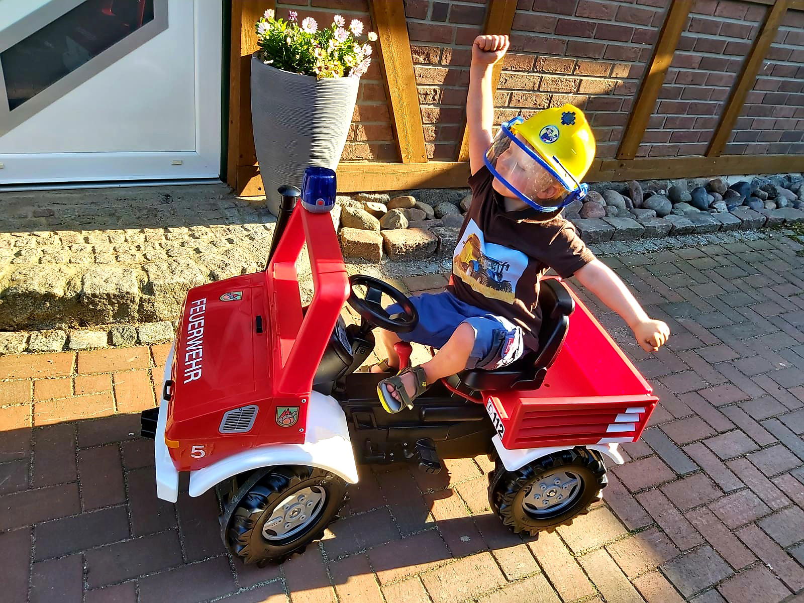 Ein kleiner Junge mit Feuerwehr-Helm sitzt auf einem kleinen Spielzeug-Feuerwehrauto und reckt eine Hand freudig in die Luft.