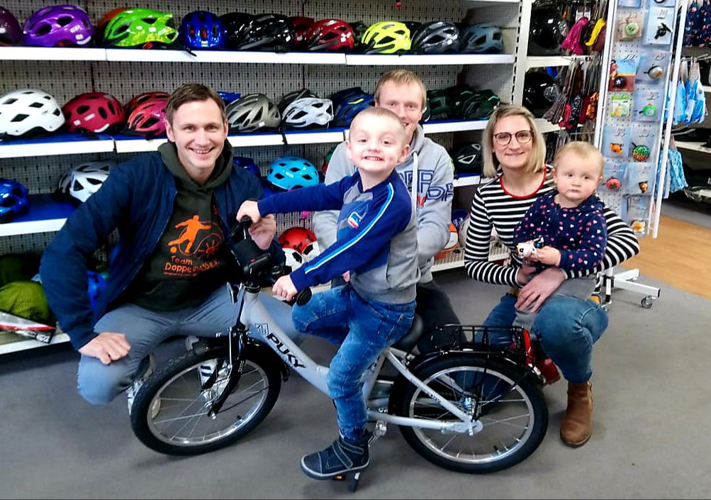 Ein Junge sitzt auf einem kleinen Fahrrad und schaut in die Kamera. Daneben knien drei Erwachsene und ein kleines Mädchen.