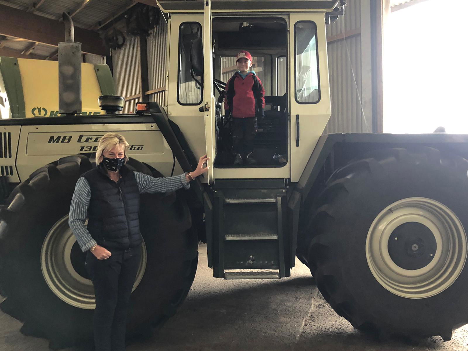 Ein Junge steht in einem Traktor und schaut in die Kamera. Eine Frau mit Maske steht ebenfalls am Trecker und schaut in die Kamera.