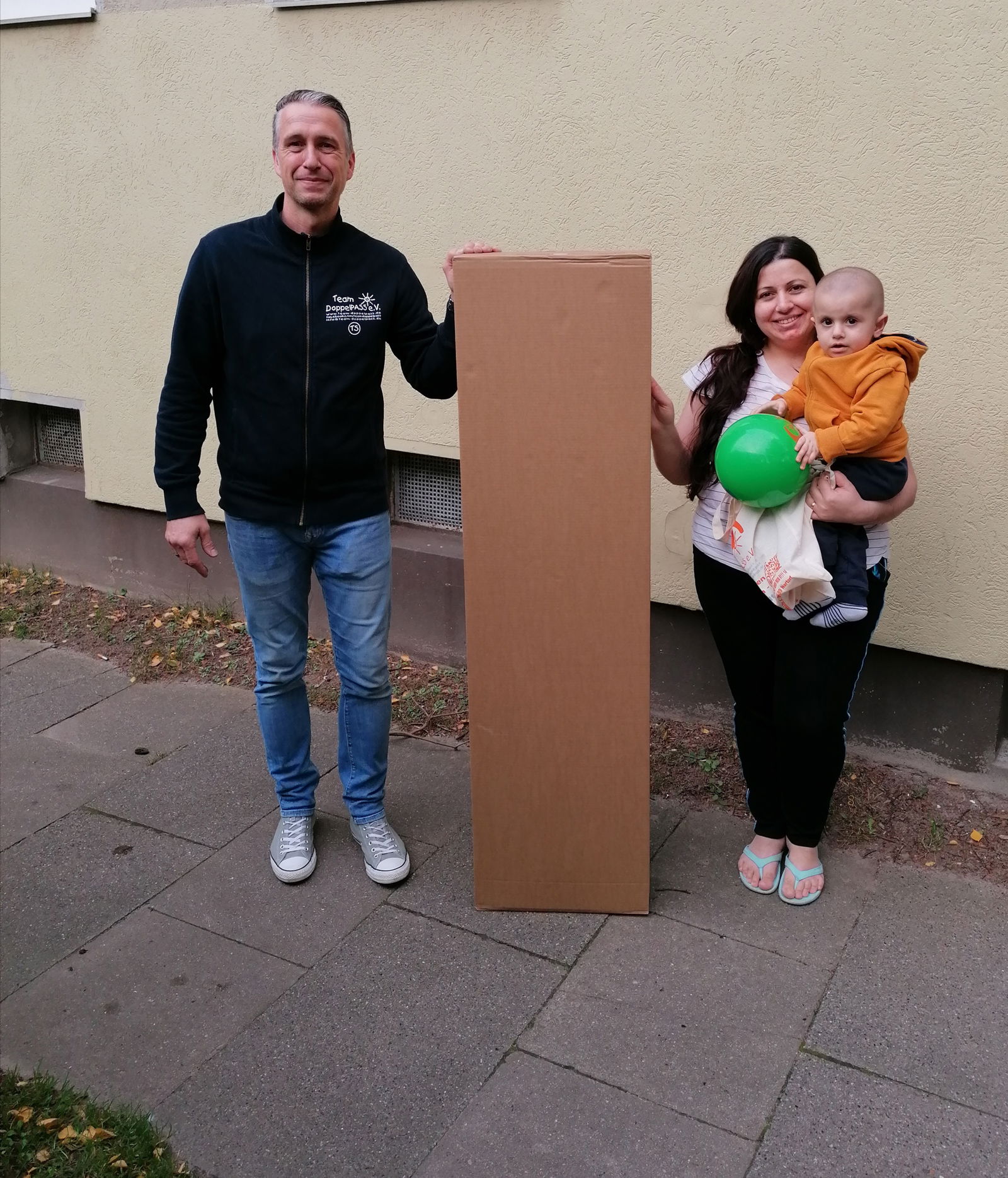 Ein Mann und eine Frau mit Kind auf dem Arm lächeln in die Kamera. Zwischen ihnen steht ein großes Paket.