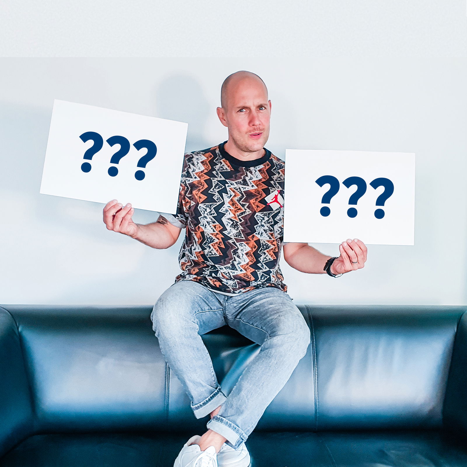 Ein Mann sitzt auf der Lehne eines Sofas und hält mit fragendem blick zwei Schilder in die Kamera, auf denen jeweils drei Fragezeichen sind.
