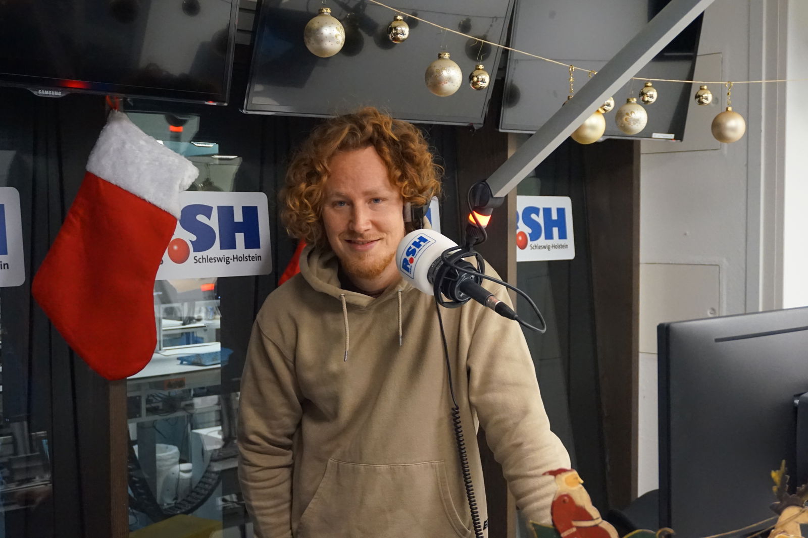 Ein rot-gelockter Mann steht in einem Radio-Studio an einem R.SH-Mikrofon und lächelt in die Kamera.