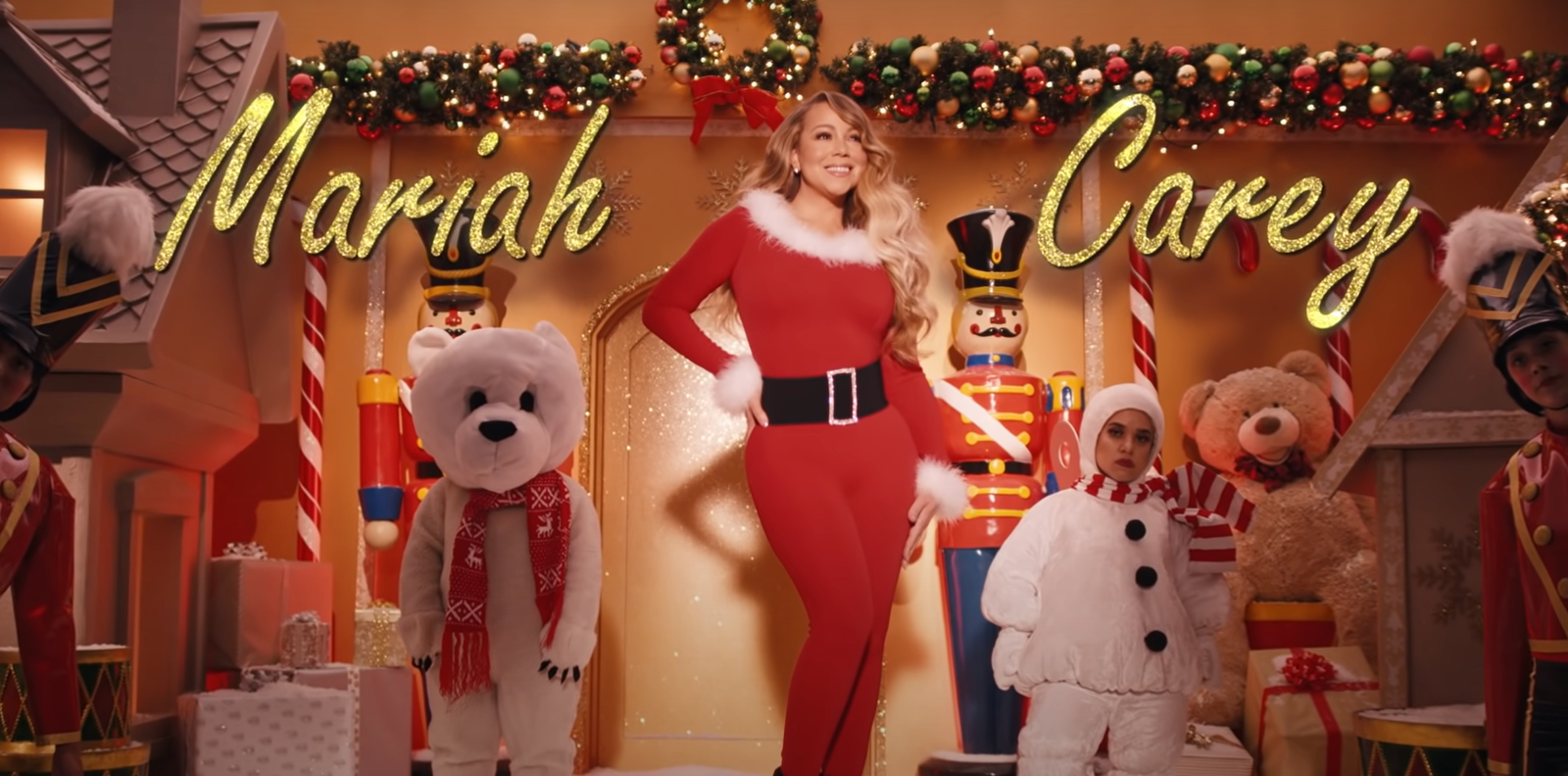 Eine weihnachtlich angezogene Frau steht in einem weihnachtlich geschmückten Zimmer, neben ihr der Schriftzug "Mariah Carey".