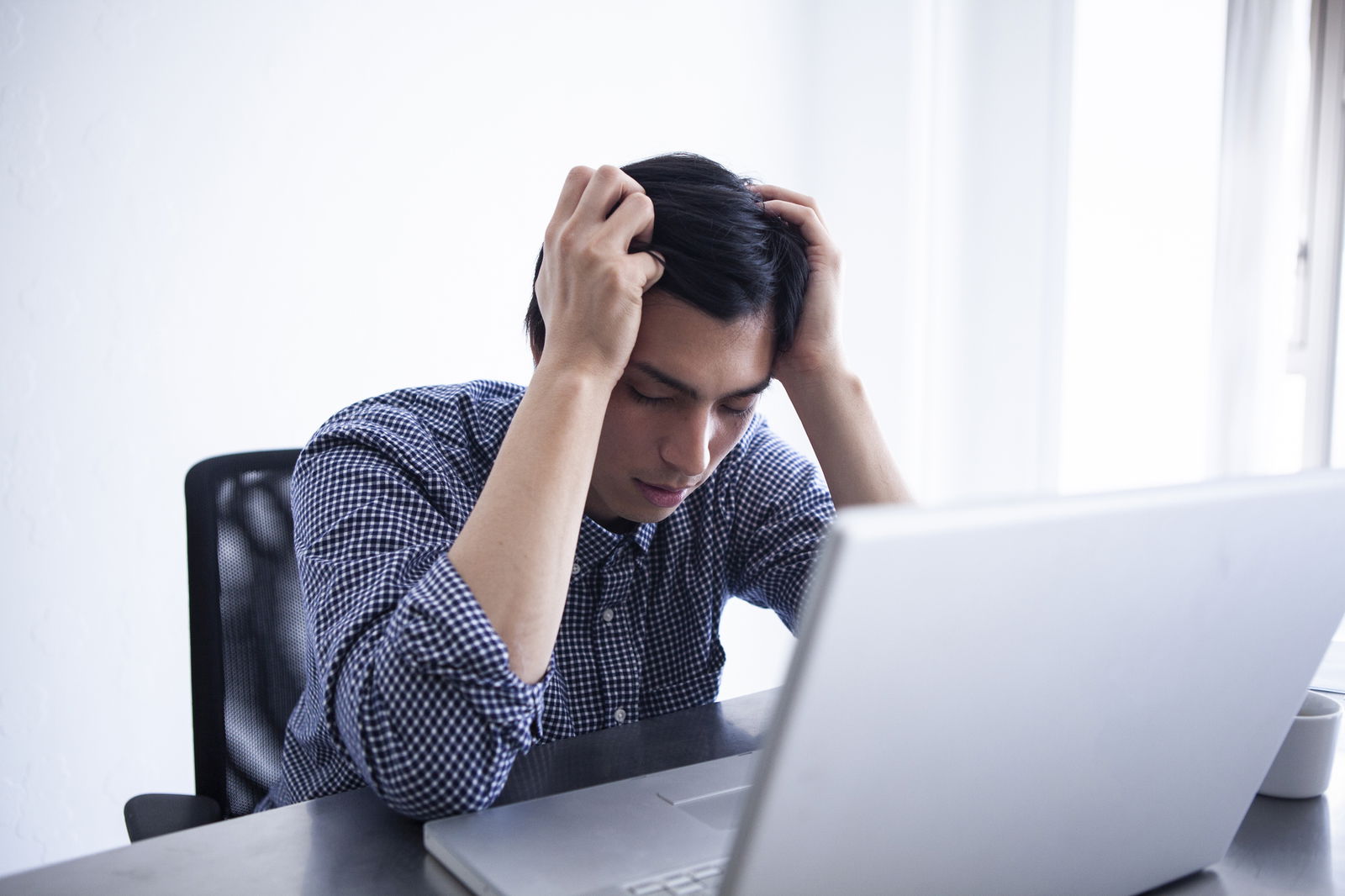 Ein junger Mann sitzt auf einem Bürostuhl vor einem Laptop und fasst sich mit beiden Händen an den Kopf.