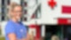 Eine blonde Pflegekraft mit Brille steht vor einem Krankenhaus und lächelt in die Kamera
