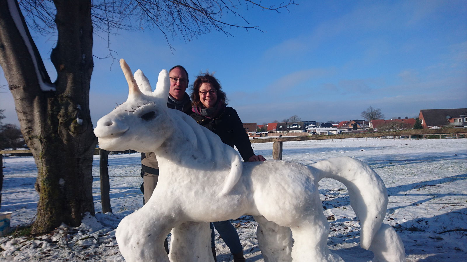 Ein Mann und eine Frau stehen in der Sonne hinter einem weißen Einhorn, das aus Schnee gebaut wurde.