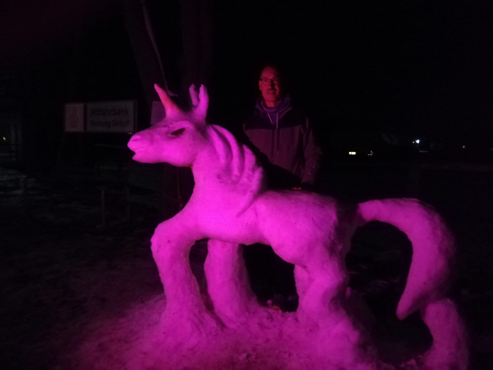 Ein Mann steht hinter einem lila beleuchteten Einhorn, das aus Schnee gebaut wurde.