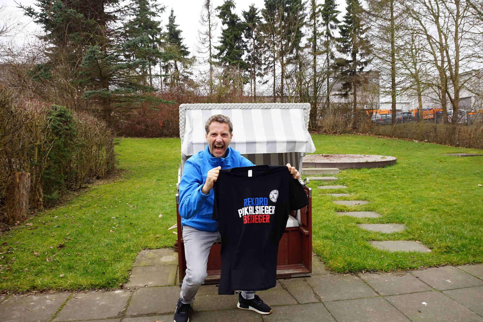 Ein Mann mit kurzen Haaren hält lächelnd ein schwarzes T-Shirt in die Kamera.