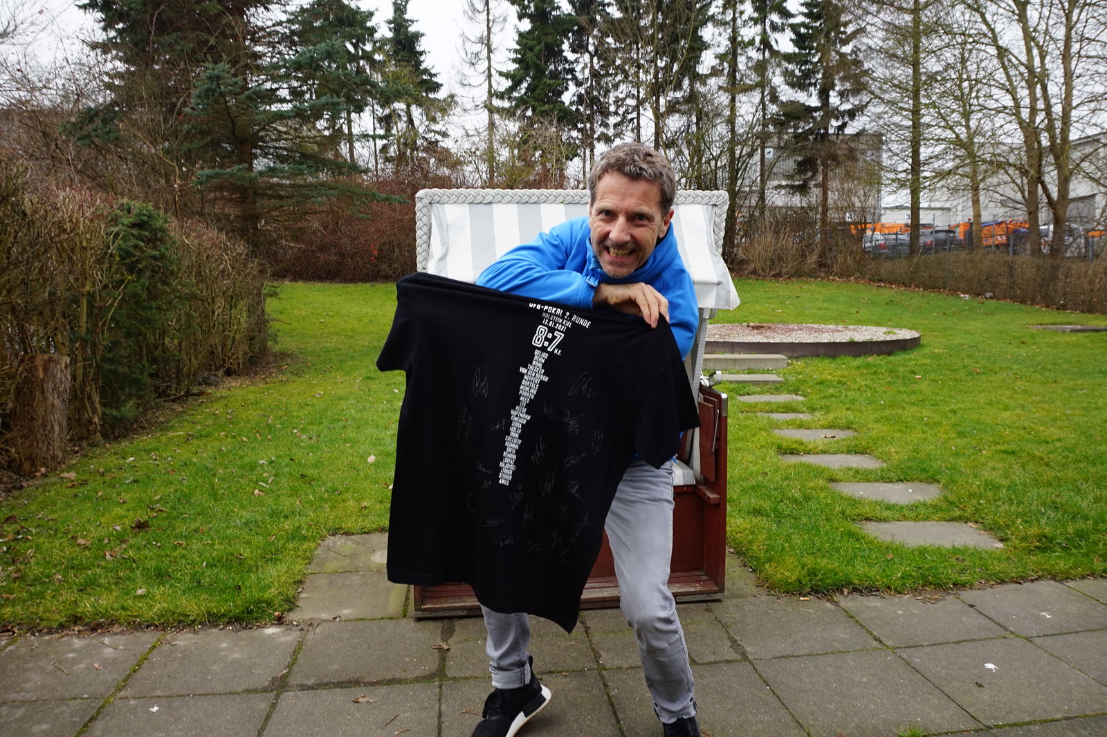 Ein Mann mit kurzen Haaren hält lächelnd ein schwarzes T-Shirt in die Kamera.