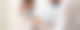 Eine Frau mit Maske in einem weißen Top sitzt. Neben ihr steht ein Mann mit einem weißen Kittel, der eine Spritze in Richtung ihres linken Oberarms hält.