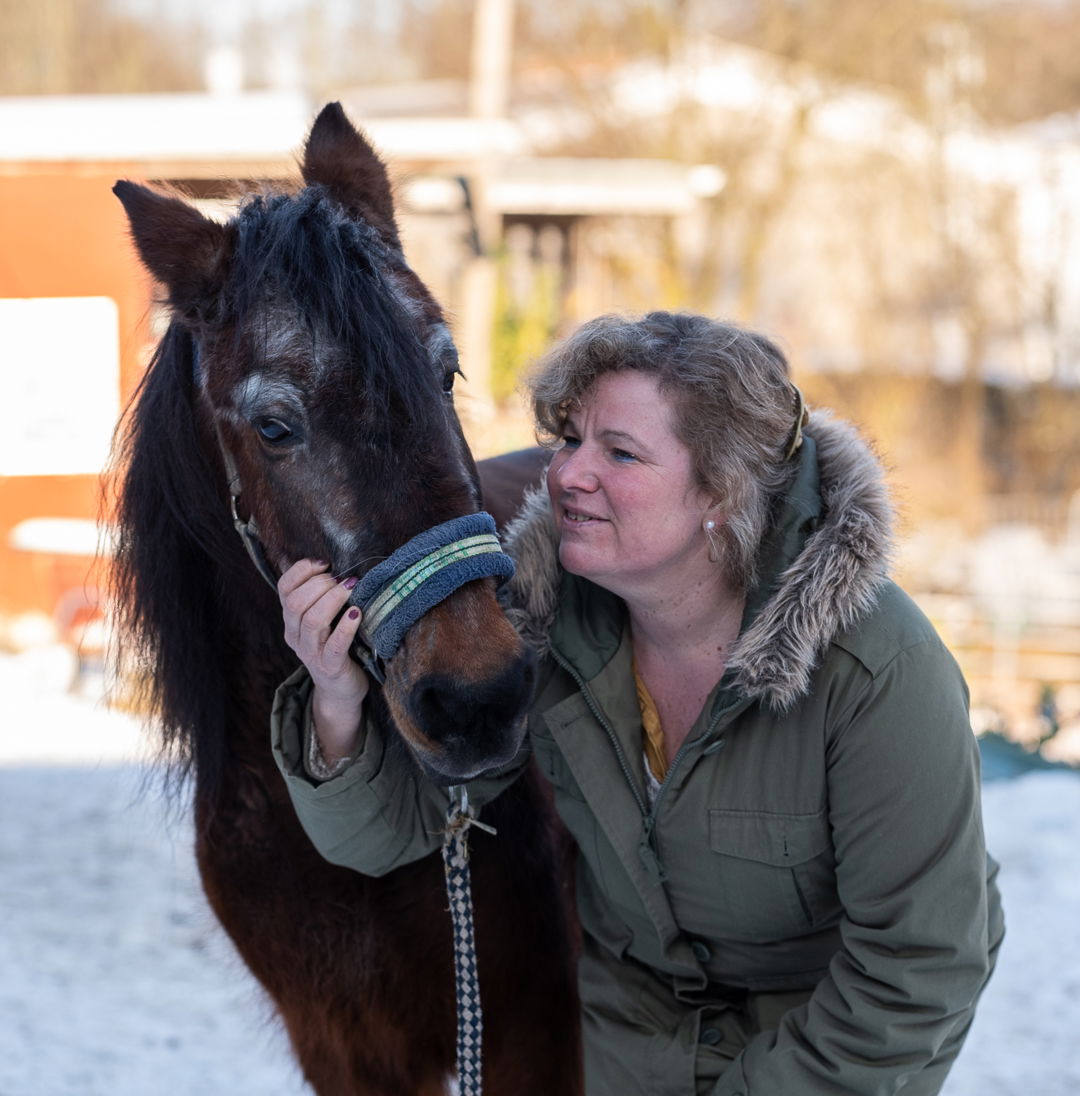 Eine Frau in Winterjacke schaut auf ein braunes Pony und hält es am Halfter
