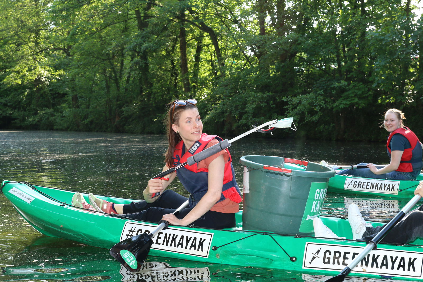 Zwei Frauen in einem grünen Kajak füllen gesammelten Müll in einen Kanister