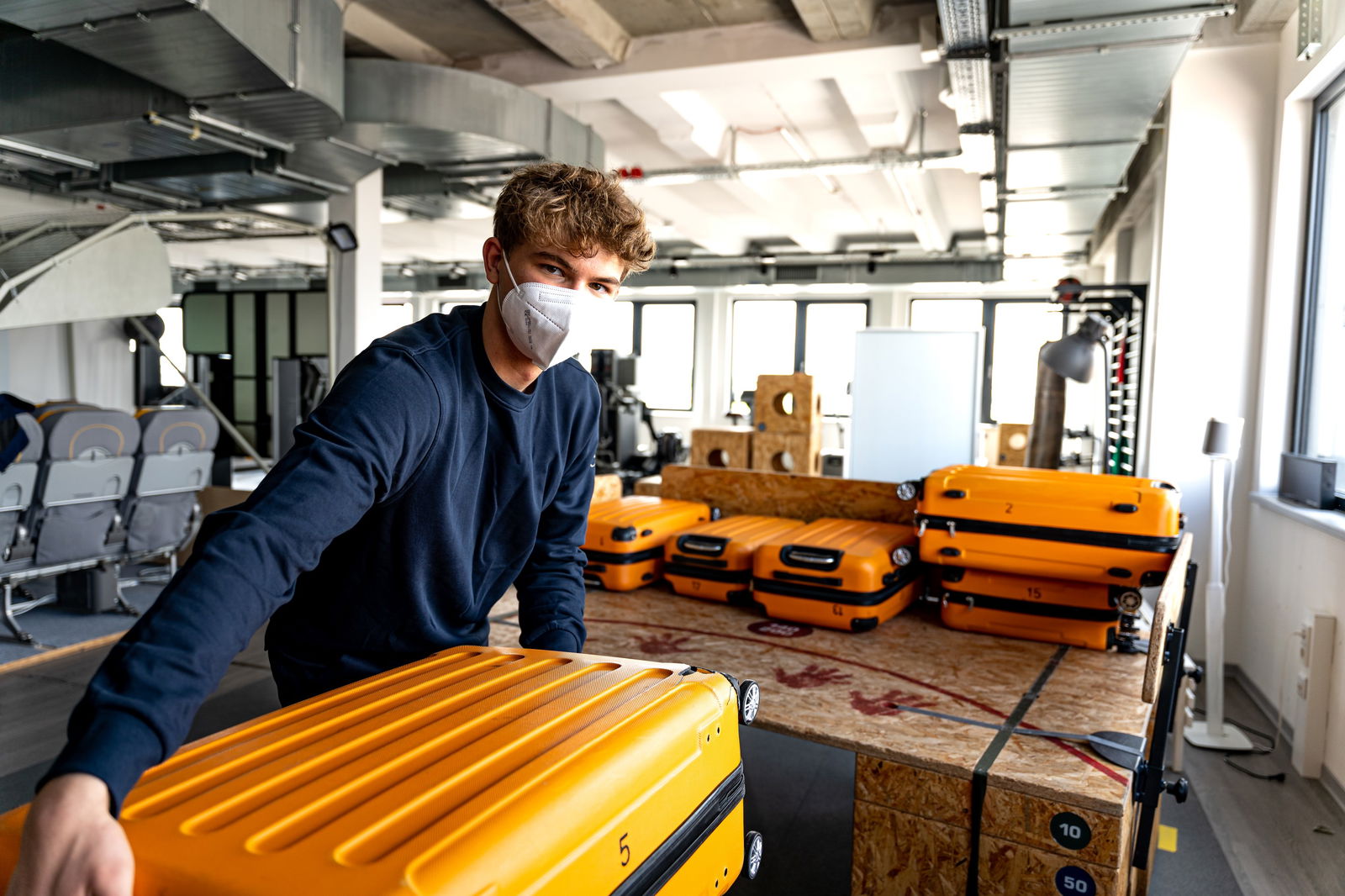 Ein junger Mann vor einem Anhänger mit Koffern hält einen orangenen Koffer in den Händen.