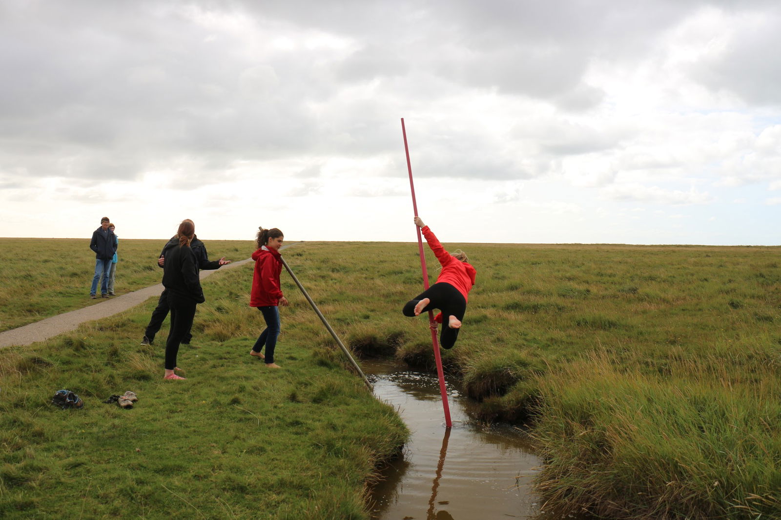 Mehrere Menschen stehen mit langen Stöcken auf einer Wiese an einem Graben und springen darüber.