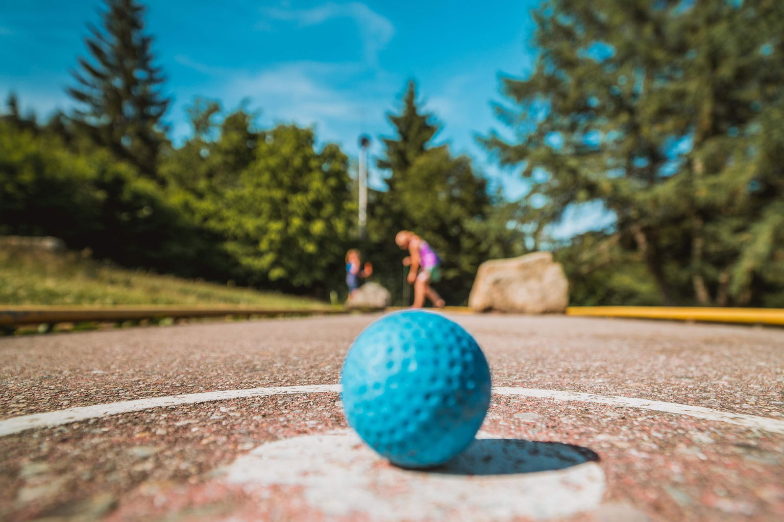 Ein blauer Golfball liegt auf einem Platz. Im Hintergrund spielen Menschen Minigolf.