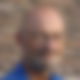Ein Mann in blauem Hemd mit Brille vor einer Mauer