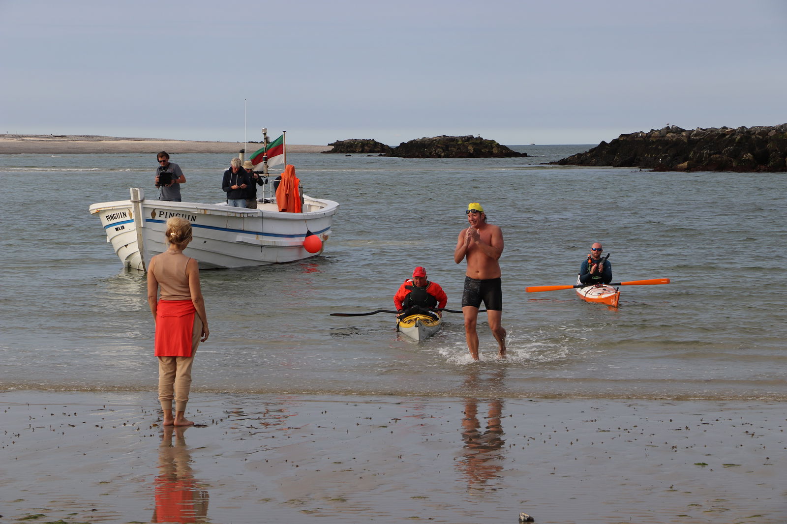 Ein Mann in Badehose steht lächelnd am Strand und eine Frau steht mit dem Rücken zur Kamera