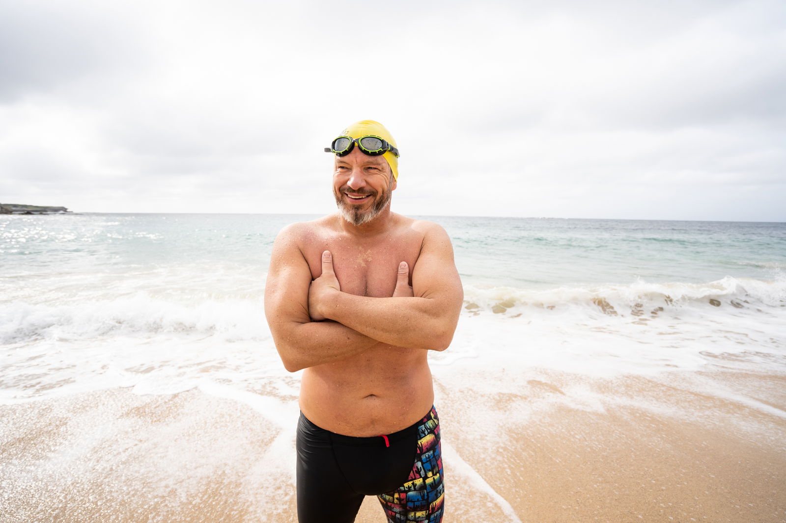 Ein Mann in Badehose und Schwimmbrille steht lächelnd am Strand und vergräbt die Arme