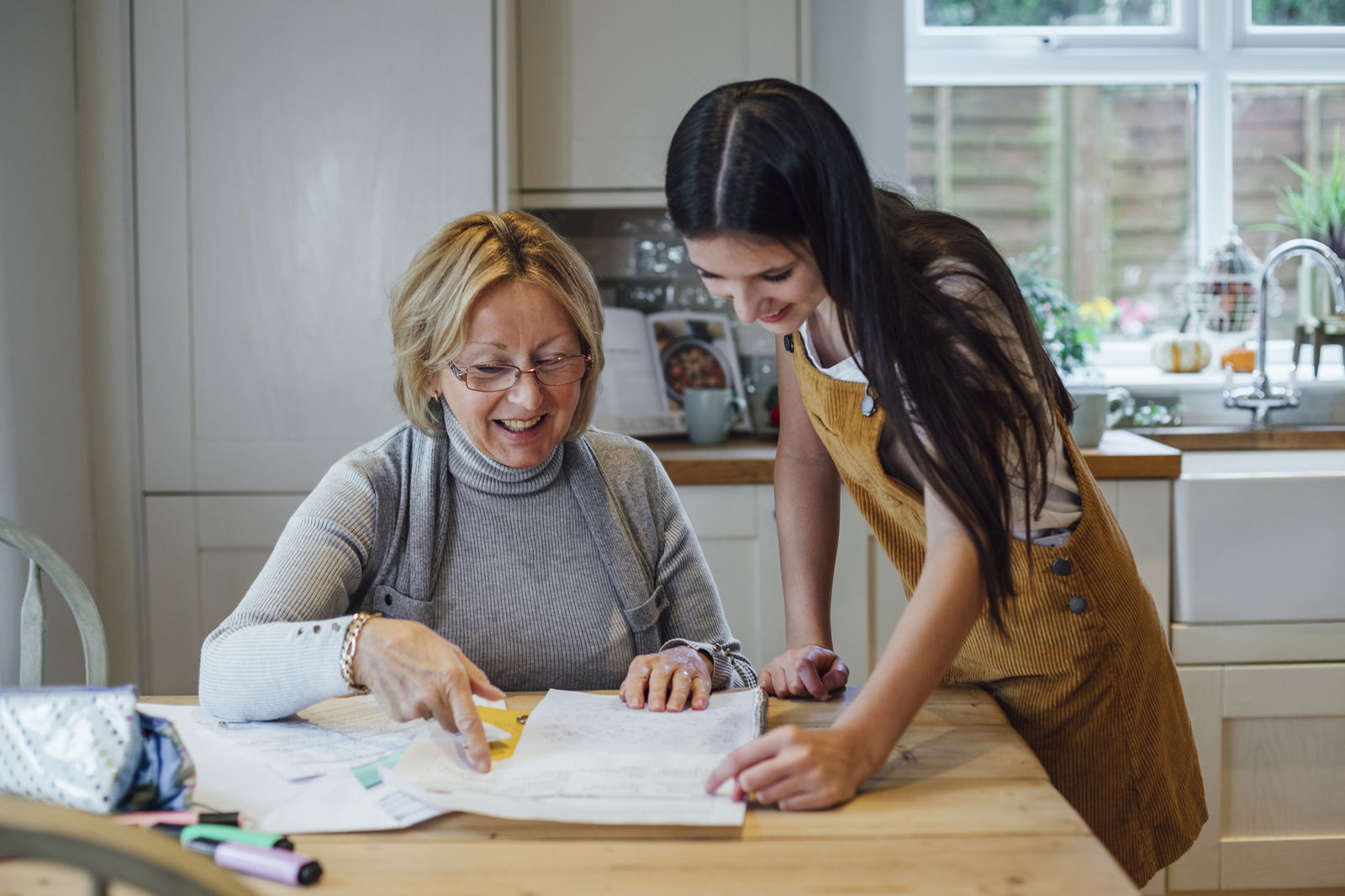 Eine ältere Dame sitzt an einem Küchentisch, Papiere vor sich. Eine junge Frau steht neben ihr am Tisch.