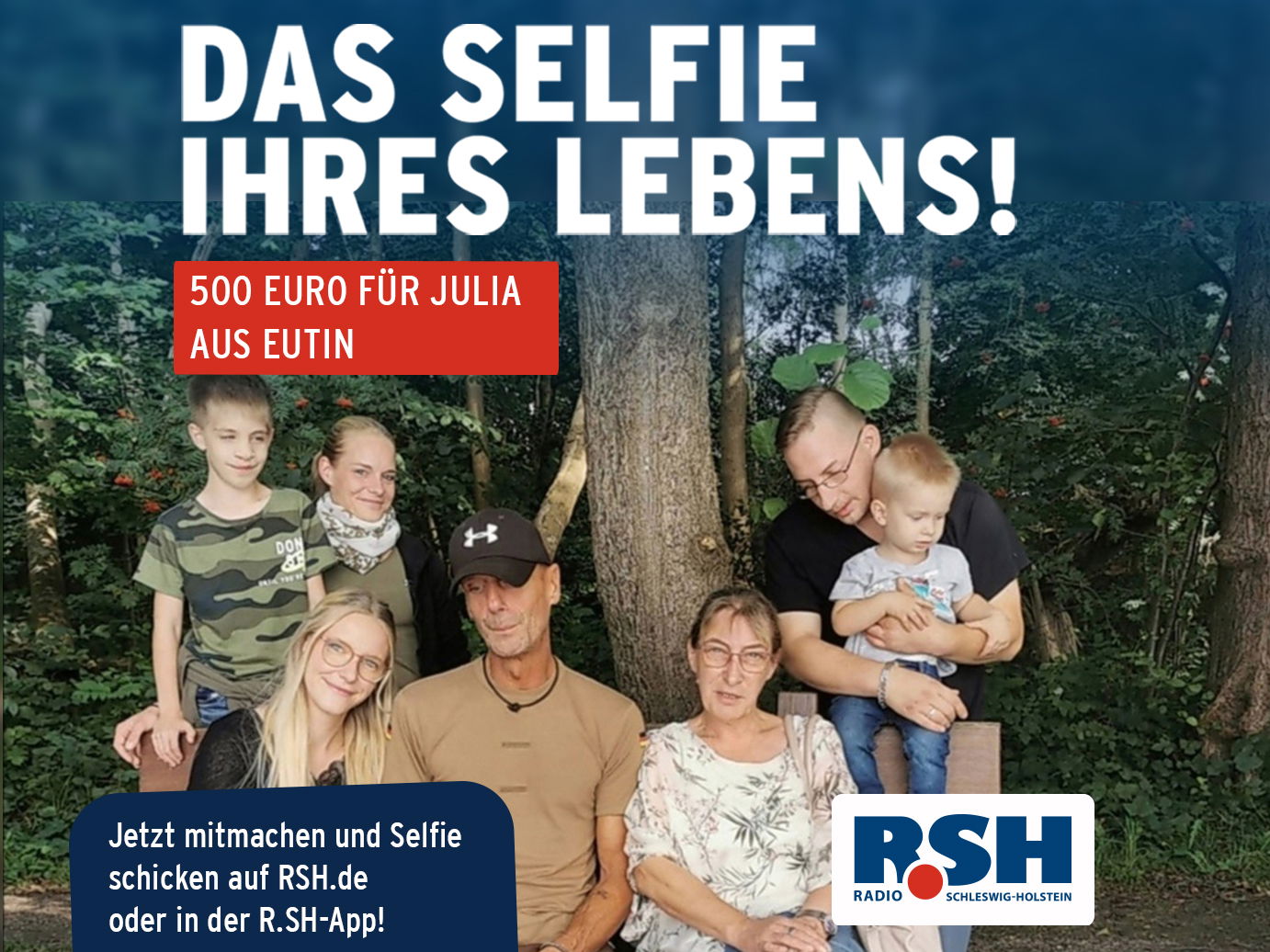 Eine Familie mit sieben Leuten sitzt vor einem Baum und lächelt in die Kamera