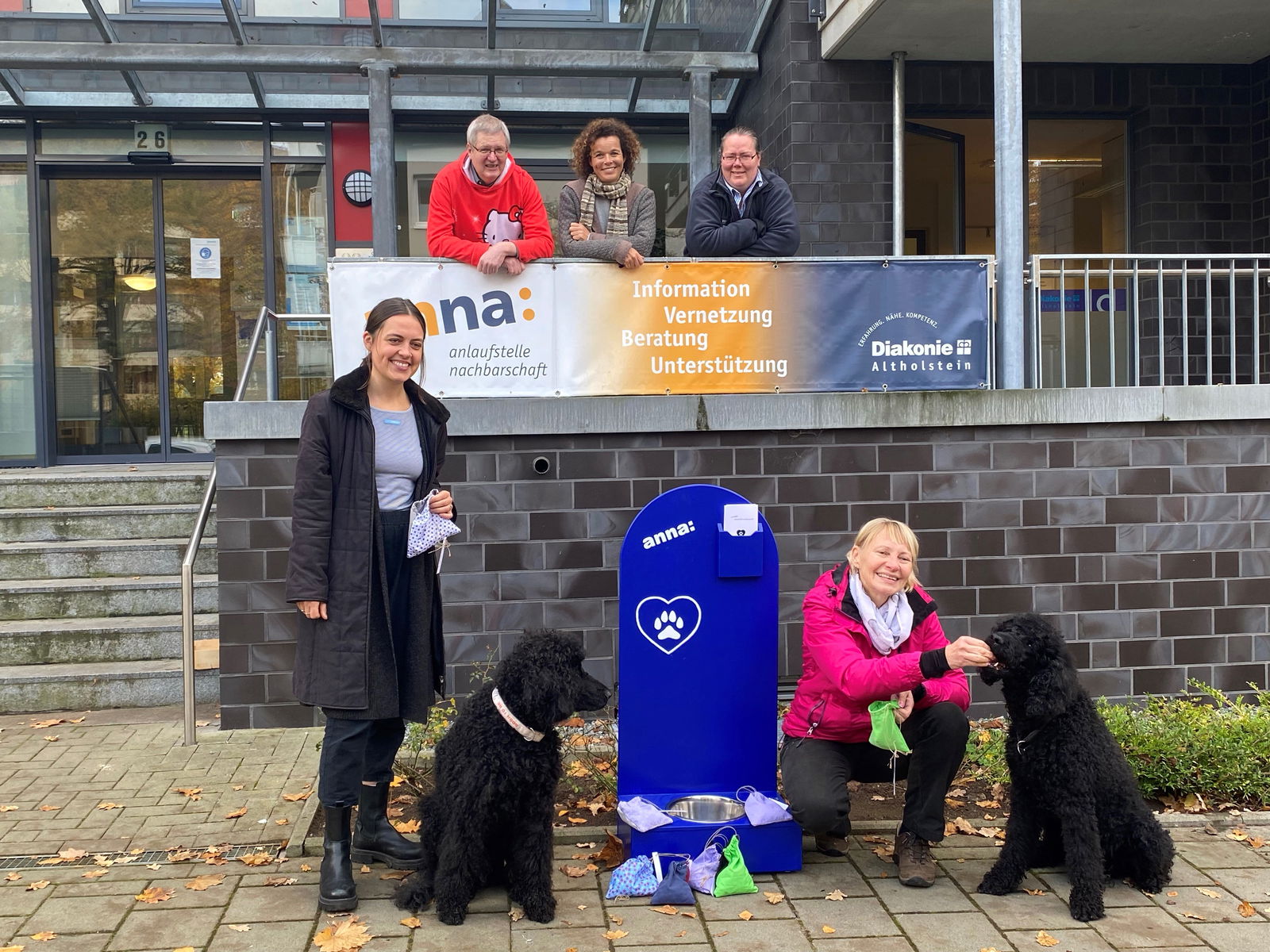 Im Vordergrund sind zwei schwarze Hund und zwei Frauen. Im Hintergrund stehen drei Personen.