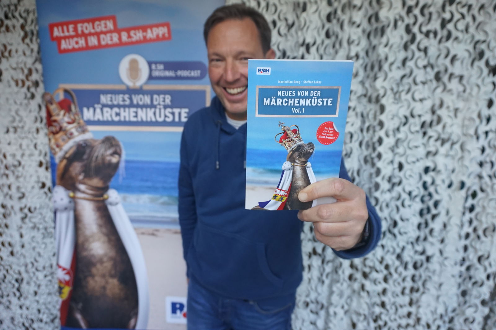 Frank Bremser steht vor einem Banner mit dem Logo von "Neues von der Märchenküste" und hält lachend ein Buch in die Kamera.