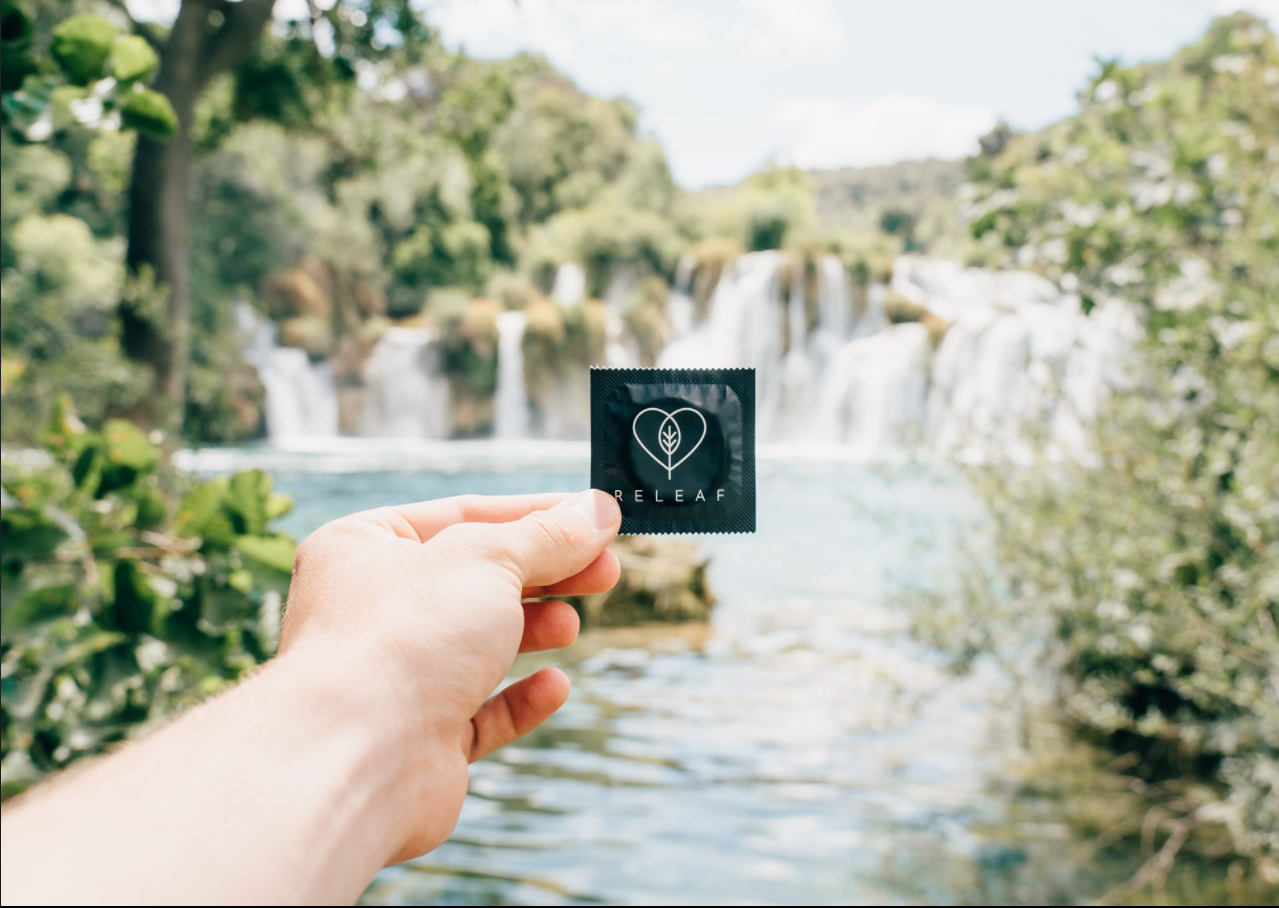 Vor einem Wasserfall hält eine Hand ein Kondom mit der Beschriftung "Releaf" in die Kamera.