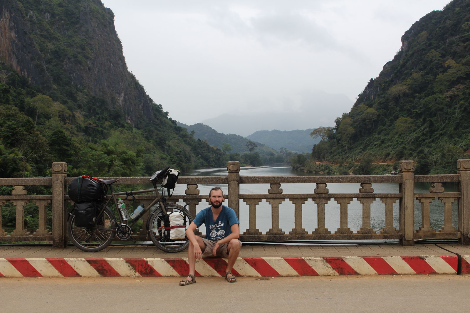 Ein Mann sitzt neben seinem Fahrrad auf einer Brücke, im Hintergrund sind Berge zu sehen.