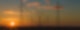 Die Sonne geht an einem leicht bewölkten Himmel unter. Davor stehen Hochspannungsmasten, an denen Kabel miteinander verbunden sind.