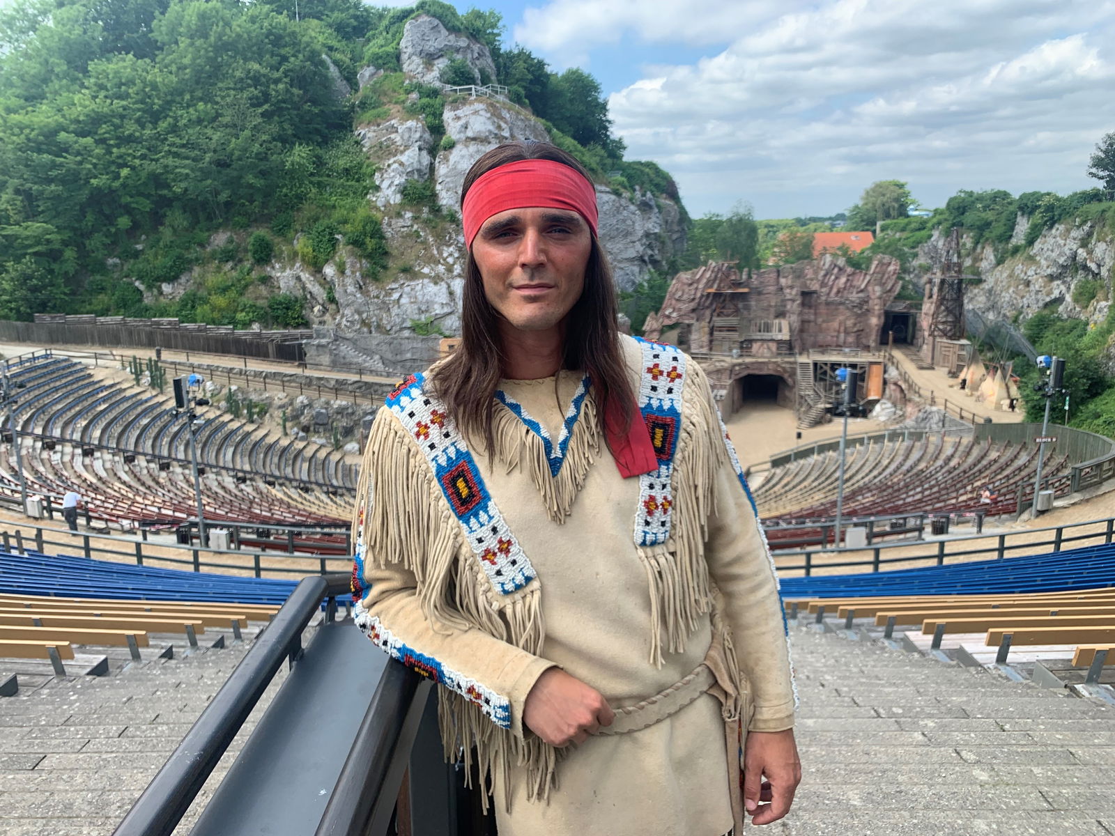 Ein junger Mann im Indianer-Kostüm steht vor einer leeren Arena und schaut direkt in die Kamera.