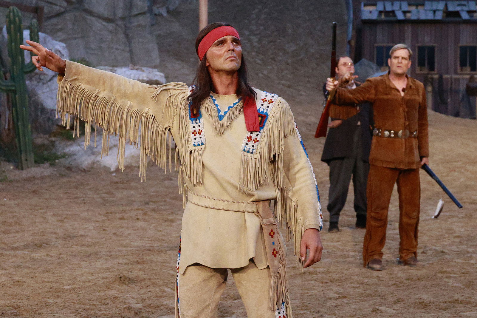 Ein Mann als Indianer steht in einer Arena. Im Hintergrund sind weitere kostümierte Darsteller zu sehen.
