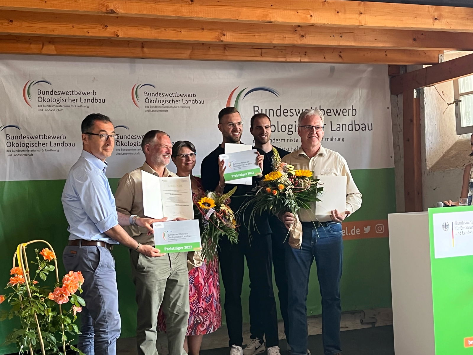 Fünf Männer und eine Frau stehen mit Urkunden und Blumensträußen auf einer Bühne und schauen lachend ins Publikum.