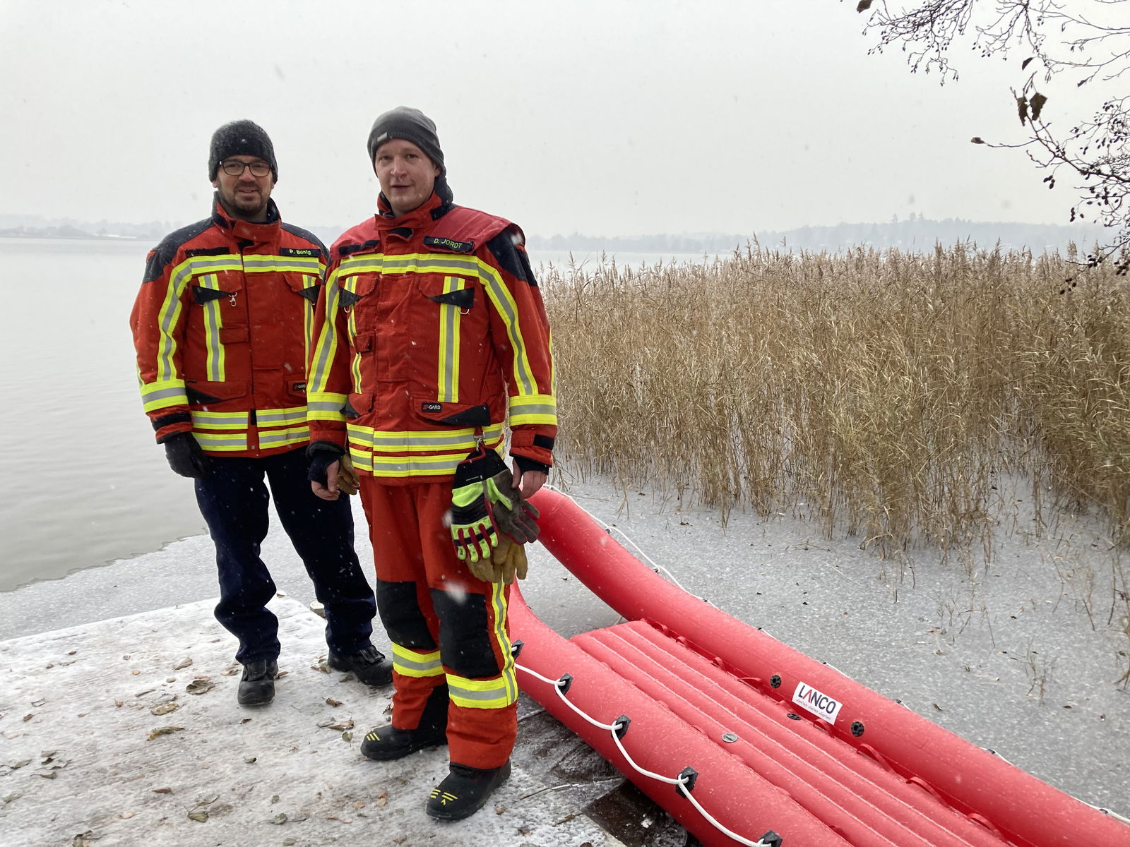 Zwei Feuerwehrmänner in roten Uniformen stehen mit einem roten Rettungsboot vor einem See.