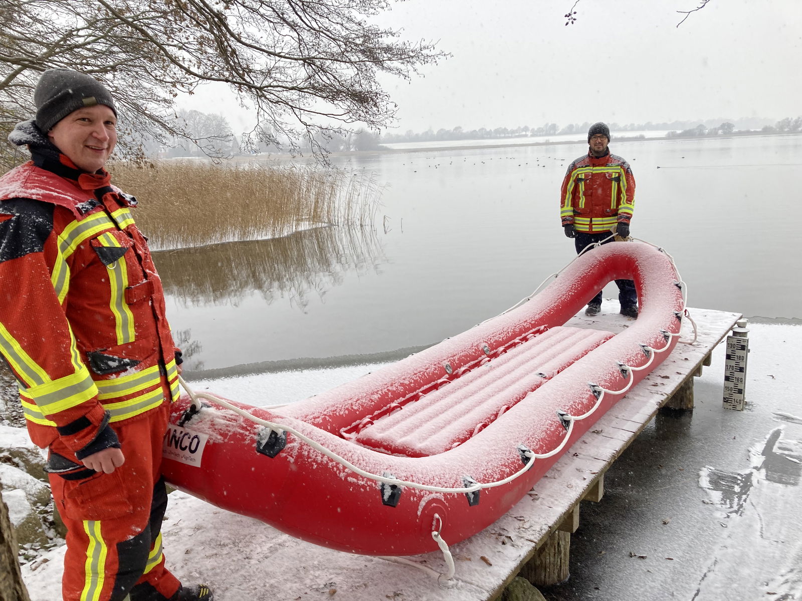 Zwei Männer in Feuerwehr-Uniformen stehen mit einer roten Matratze vor einem See.