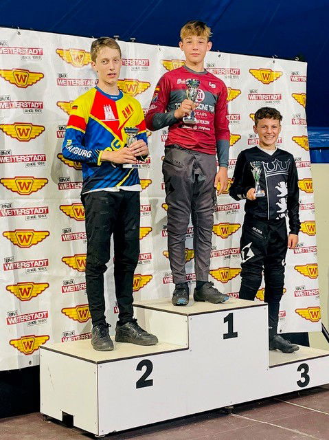 Drei Jungen in Rennanzügen auf einem Podium mit Pokalen.