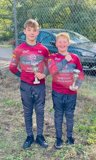 Zwei Jungen in roten Trikots lachen mit Pokalen in der Hand in die Kamera.