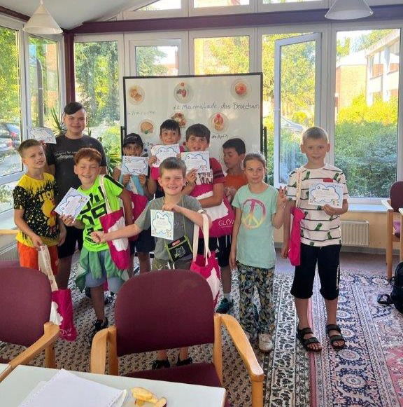 Kinder stehen mit selbstgemalten Bildern vor einer Tafel und lachen in die Kamera.