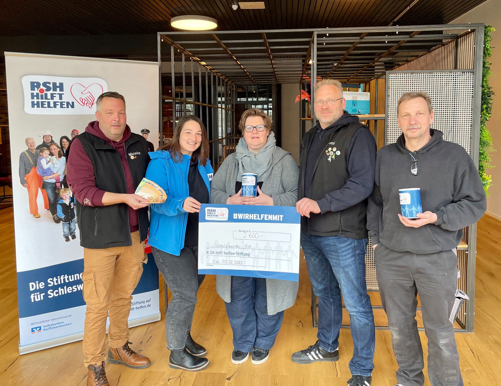 Drei Männer und zwei Frauen stehen mit einem Scheck und Spendendosen vor einem Banner mit der Aufschrift "R.SH hilft helfen".
